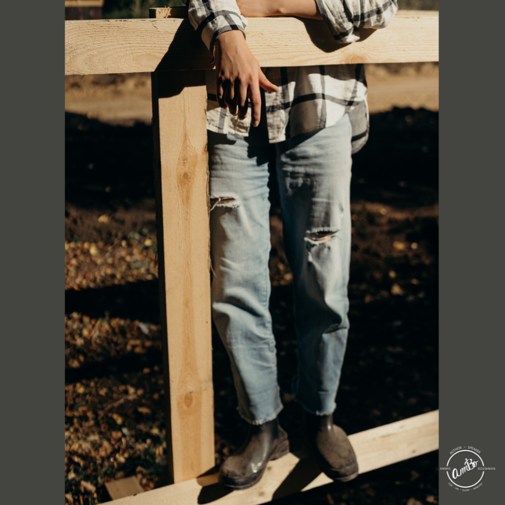 A man in jeans and boots leaning on a wooden fence.