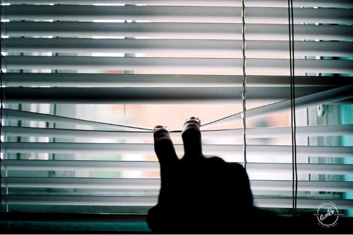 A person sitting on the ground with their feet up looking out of blinds.